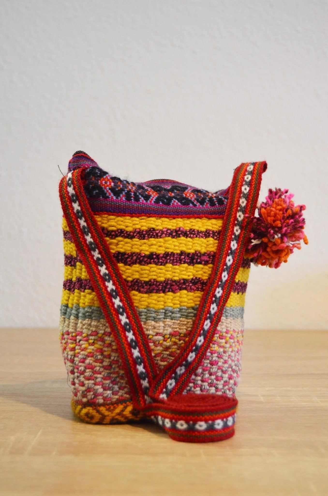 Handmade small shoulder bag with colorful alpaca knit and red patterned strap with pom-pom detail.