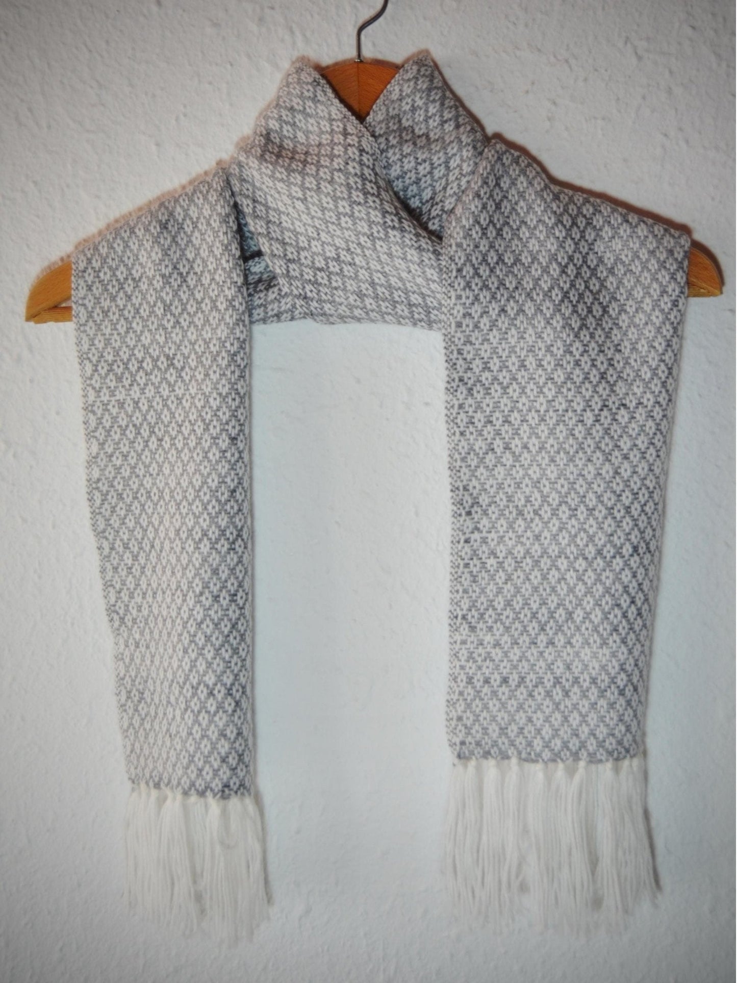 Handmade light gray alpaca shawl with diamond pattern and fringed ends hanging on wooden hanger.